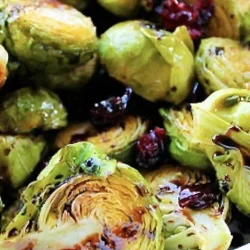 A close-up of a white dish filled with roasted Brussels sprouts, some halved and charred, mixed with cranberries and drizzled with rich balsamic reduction.
