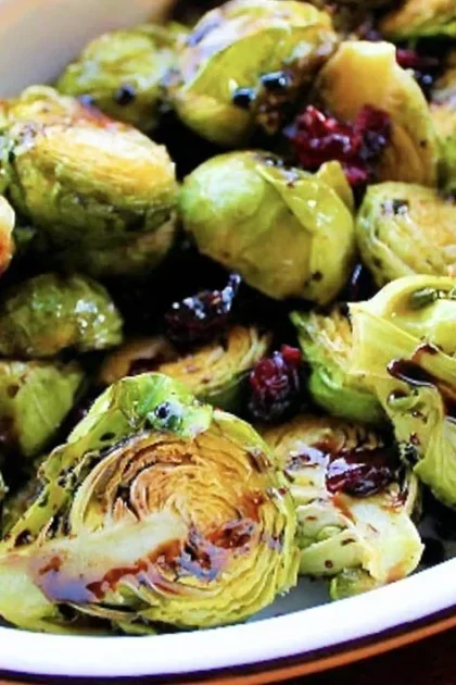 A close-up of a white dish filled with roasted Brussels sprouts, some halved and charred, mixed with cranberries and drizzled with rich balsamic reduction.