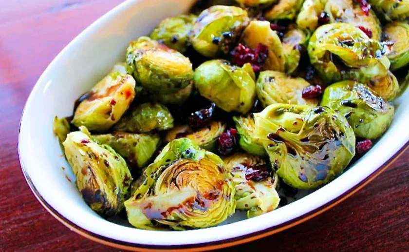A white oval dish filled with roasted Brussels sprouts, garnished with cranberries and a drizzle of balsamic reduction, sits on a wooden surface.