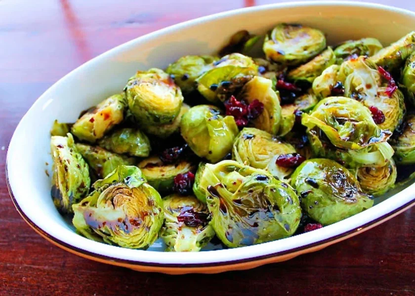 A white oval dish filled with roasted Brussels sprouts, some caramelized, garnished with cranberries and drizzled with a rich balsamic reduction. The dish is placed on a wooden surface.