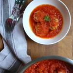 Un tazón de albóndigas de lomo de cerdo en salsa de tomate, adornadas con perejil, se encuentra en una mesa de madera junto a una sartén con más albóndigas, tenedores, cuchillos y una servilleta de tela a rayas.