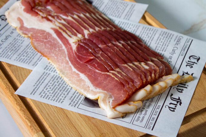 A slack of sliced bacon before being cooked. The bacon is placed over deli paper that simulates a newspaper.
