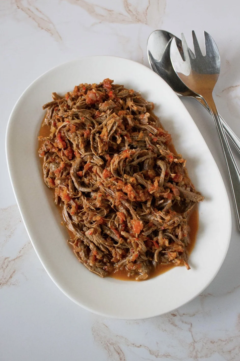 Carne mechada venezolana desmenuzada con salsa roja servida en un plato blanco ovalado, con utensilios para servir colocados al lado del plato sobre una superficie de mármol.