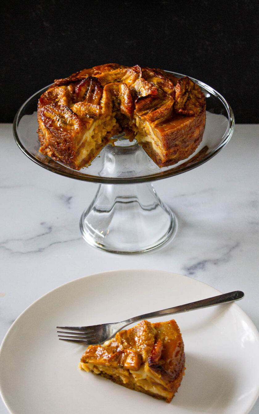 Torta de platano served on a glass cake dish, plus a slice of the cake serves on a white plate with a fork.