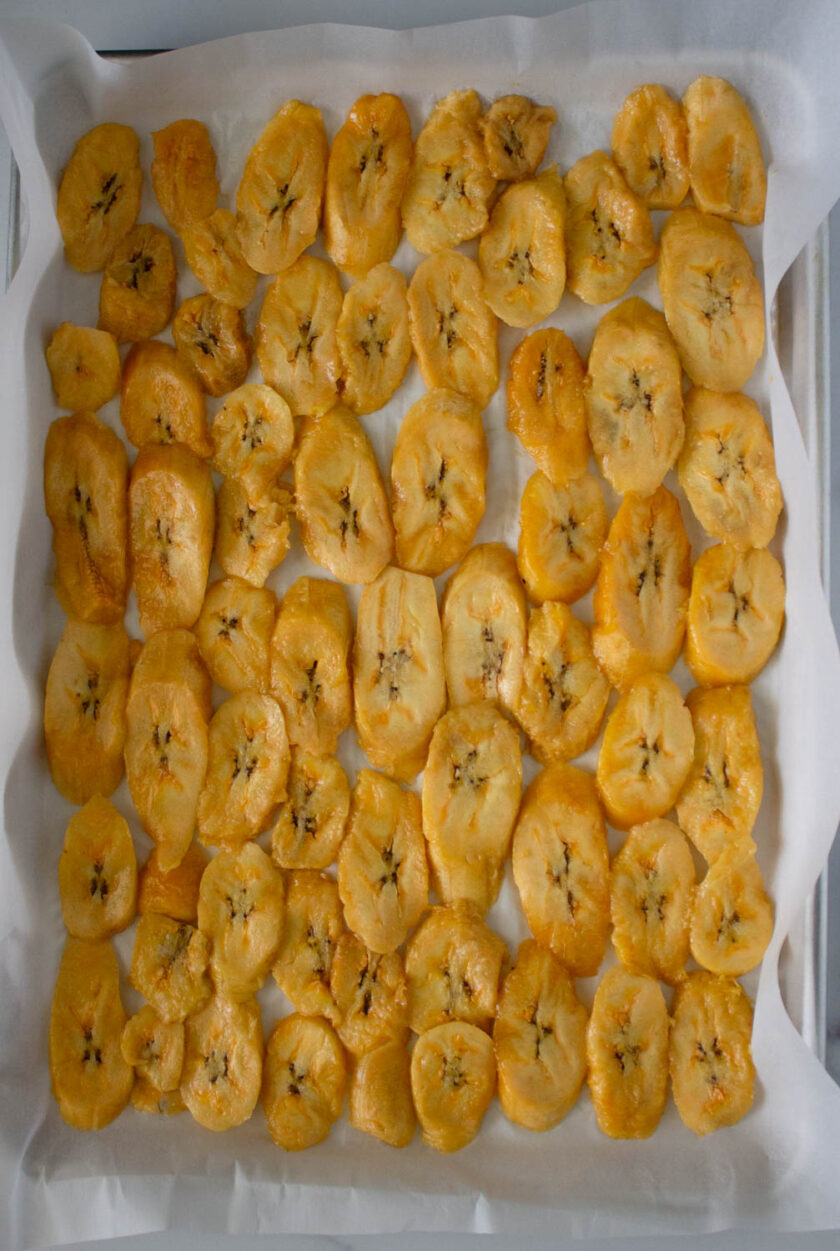 A baking sheet, covered with parchment paper, and filled with raw plantain slices.