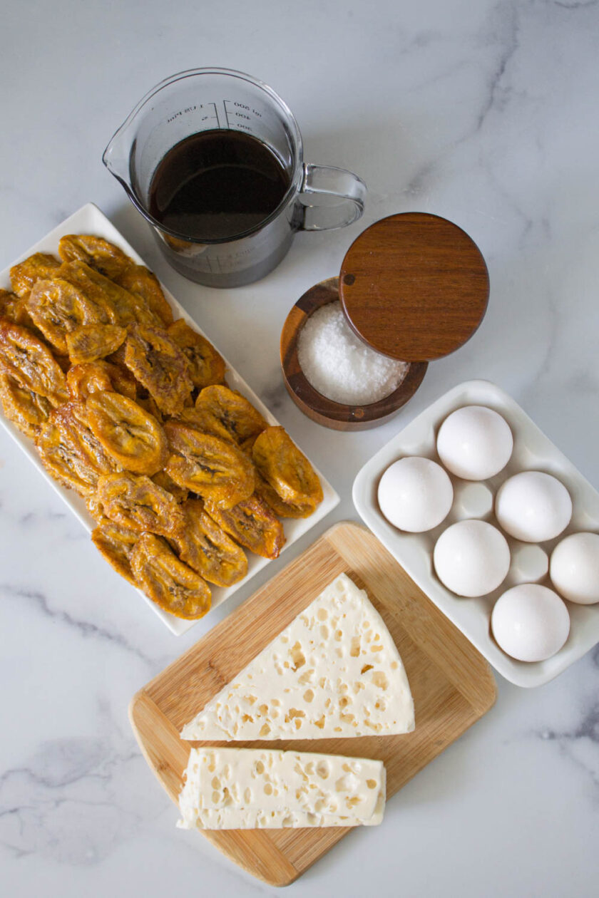 Ingredients to make Venezuelan torta de platano, including plantains, papelón syrup, salt, eggs and white cheese.
