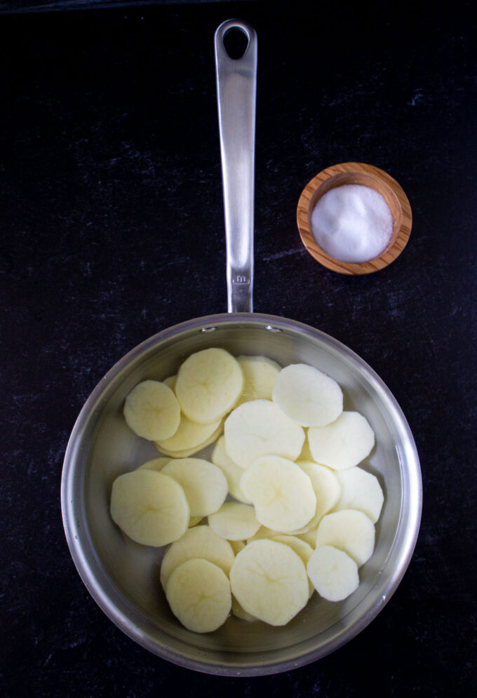 Sliced potatoes on a saucier with water, there is wooden bowl with salt, tom