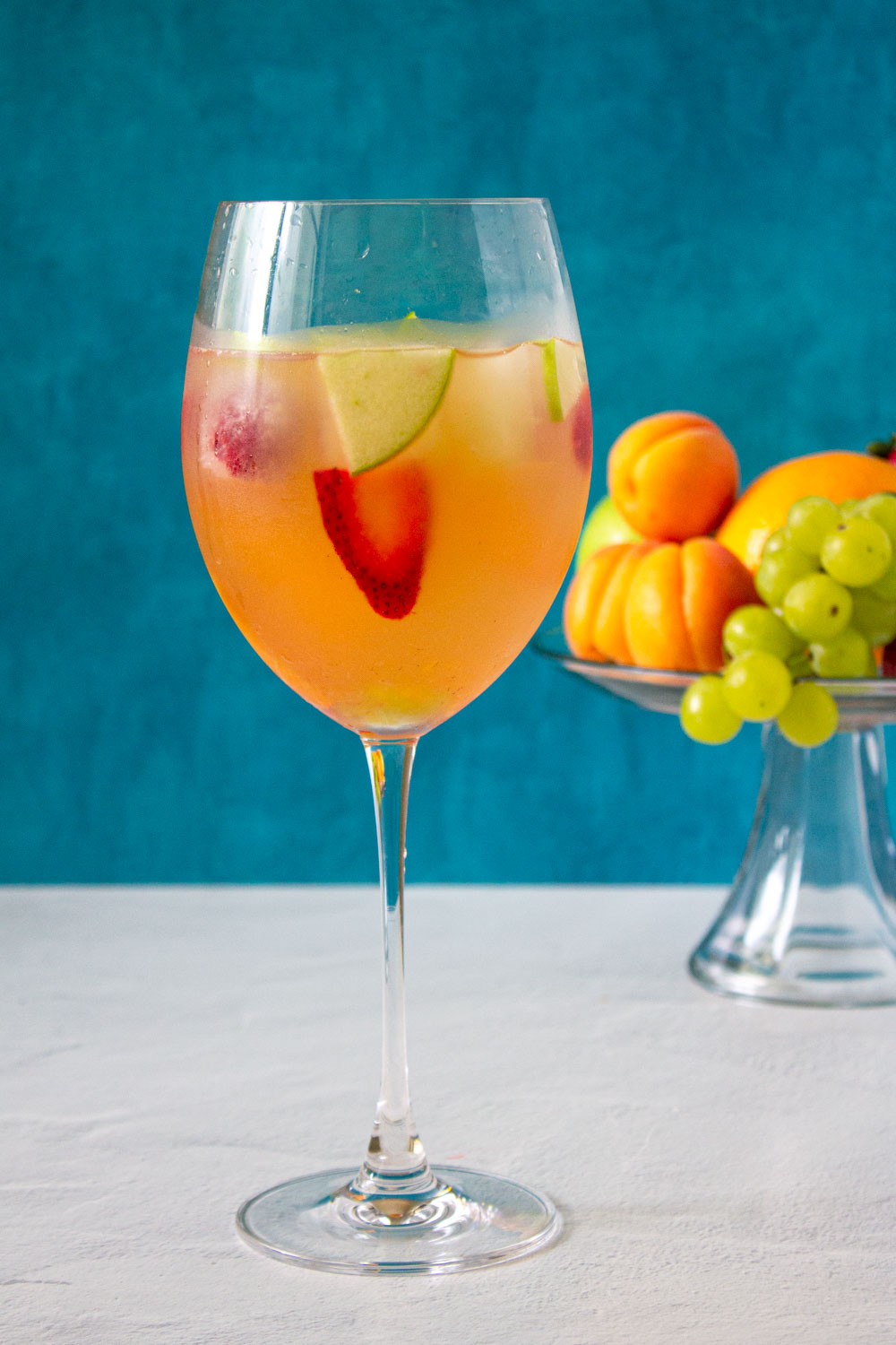 Copa de sangría de vino blanco. Al fondo se ve una jarra de sangría y también frutas frescas.