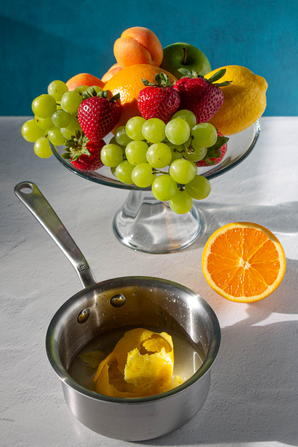 Olla pequeñas con los ingredientes para hacer jarabe de limón, y una fuente con fruta fresca, incluyendo: uvas verdes, limones, manzanas verdes, frescas, albaricoques y naranjas.