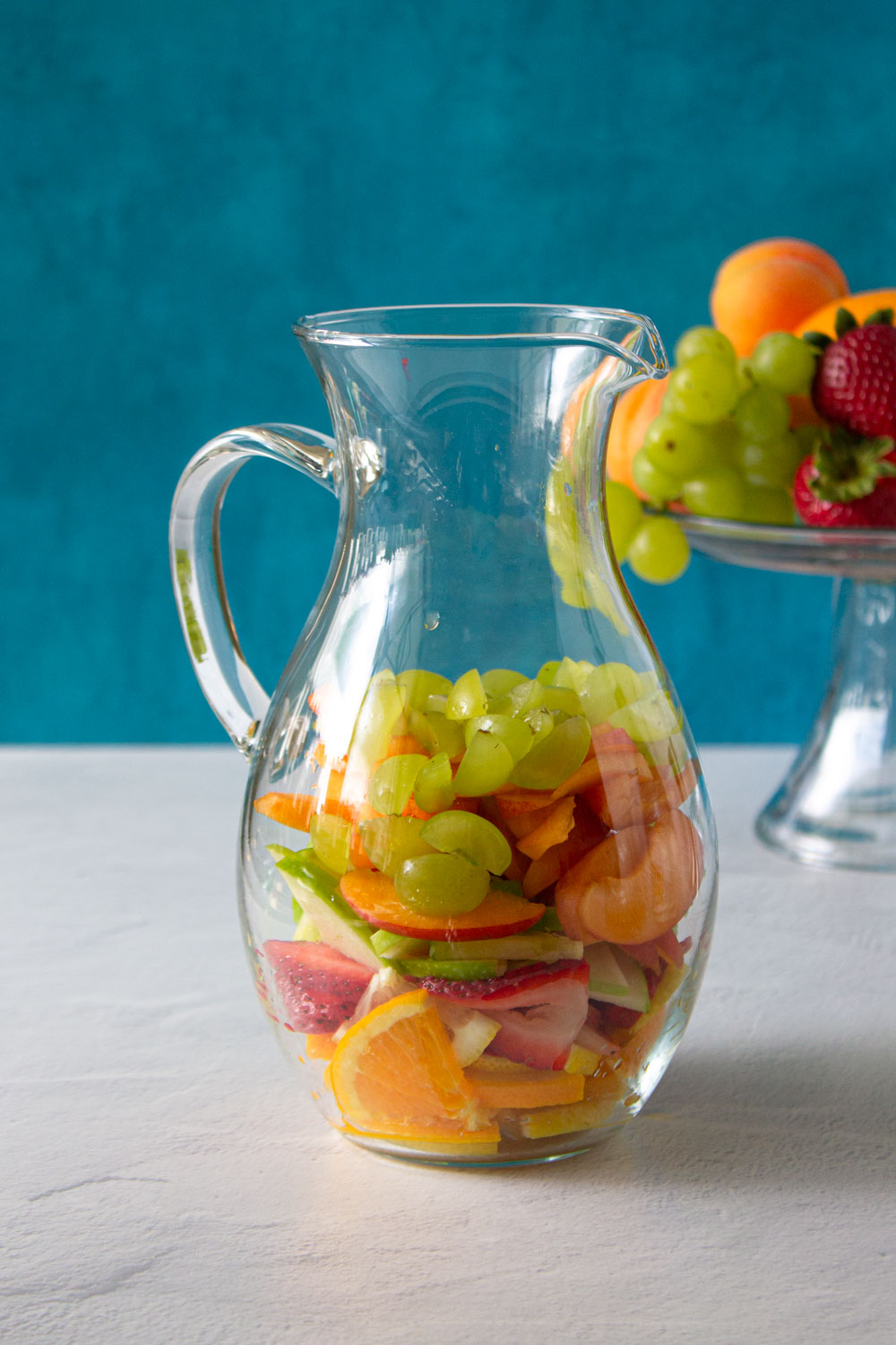 Jarra de cristal llena por la mita con frutas cortadas para hacer sangría blanca.