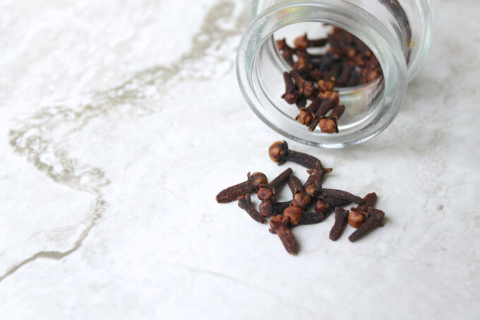 Close up of a glass jar whit whole cloves, plus a a few cloves over a marble top, by enrilemoine.