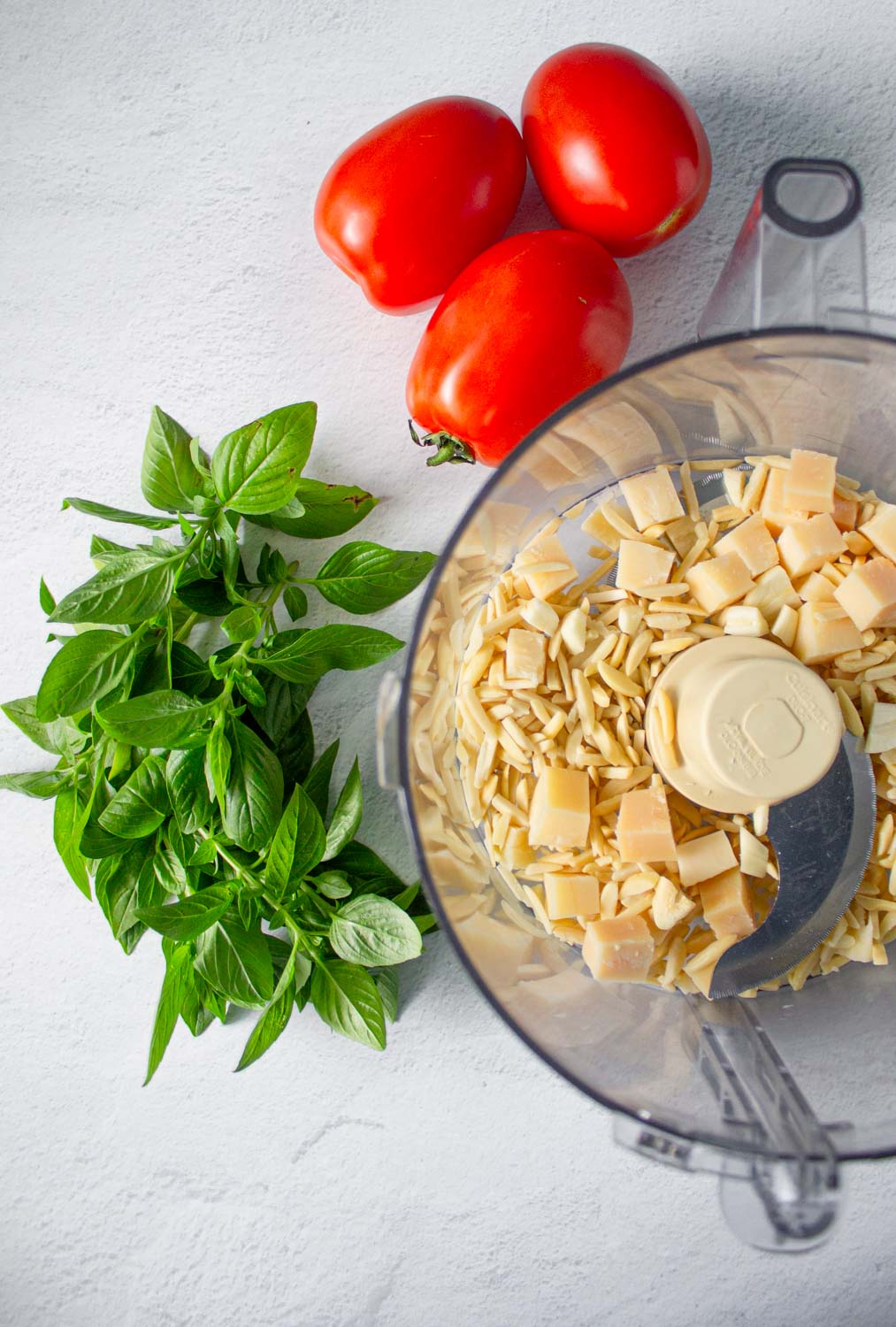 Vista cenital del tazón de un procesador de alimentos con almendras peladas y dados de queso parmesano, al lado se ven tomates y albahaca fresca, by enrilemoine.