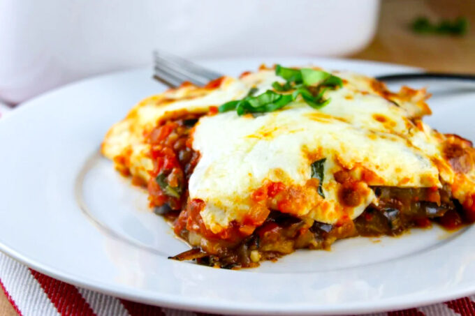 Porción de lasaña de berenjena, servida en un plato blanco, con un tenedor al fondo, by enrilemoine.