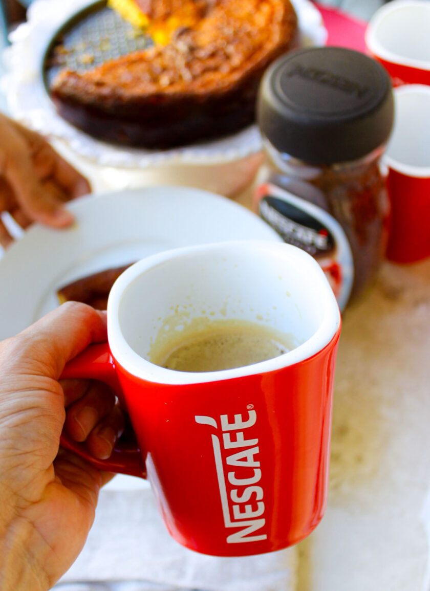 Una persona sosteniendo una taza de café junto a un plato de torta de jojoto.