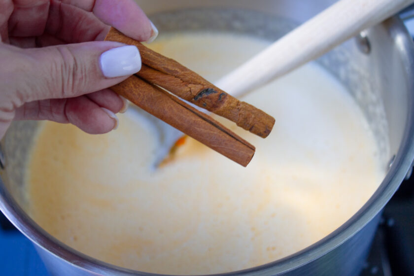 Una mano sosteniendo unas ramitas de canela en una olla de ponche crema.