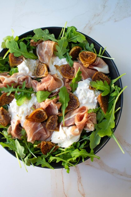 Ensalada de hugos, prosciutto, burrata y arugula en una ensaladadera negra.