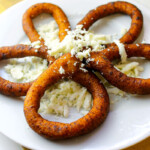 Five fried mandocas (plantain rings) rings arranged in a flower shape on a white plate, topped with crumbled white cheese.