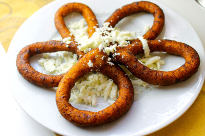 Five fried mandocas (plantain rings) rings arranged in a flower shape on a white plate, topped with crumbled white cheese.