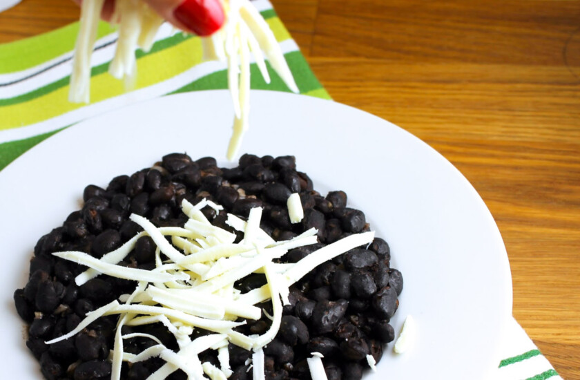 Un plato de caraotas negras refritas alque alguien le agrega queso blanco rallado.
