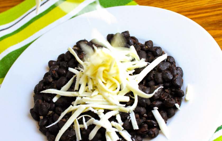 Un plato de caraotas negras refritas alque alguien le agrega queso blanco rallado.