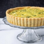A leek quiche on a glass pedestal against a marbled background.
