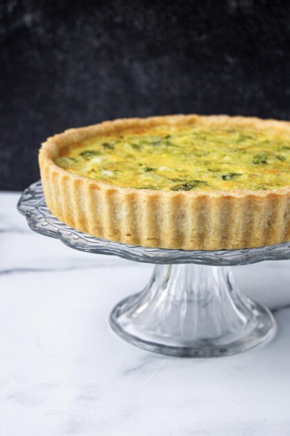 A leek quiche on a glass pedestal against a marbled background.