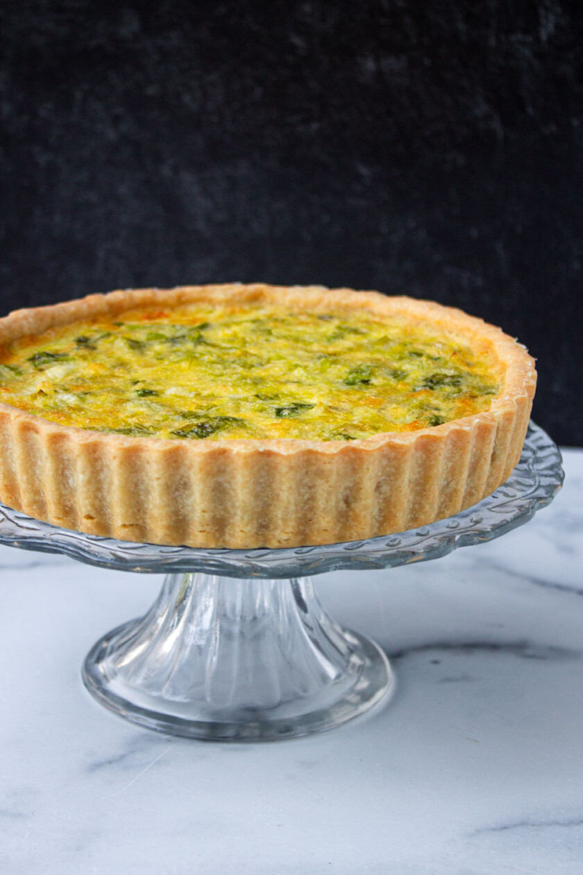 Una quiche recién horneada sobre un pedestal de cristal sobre un fondo de mármol.