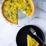 A slice of quiche on a black plate with a fork, alongside the remaining quiche on a glass serving dish.