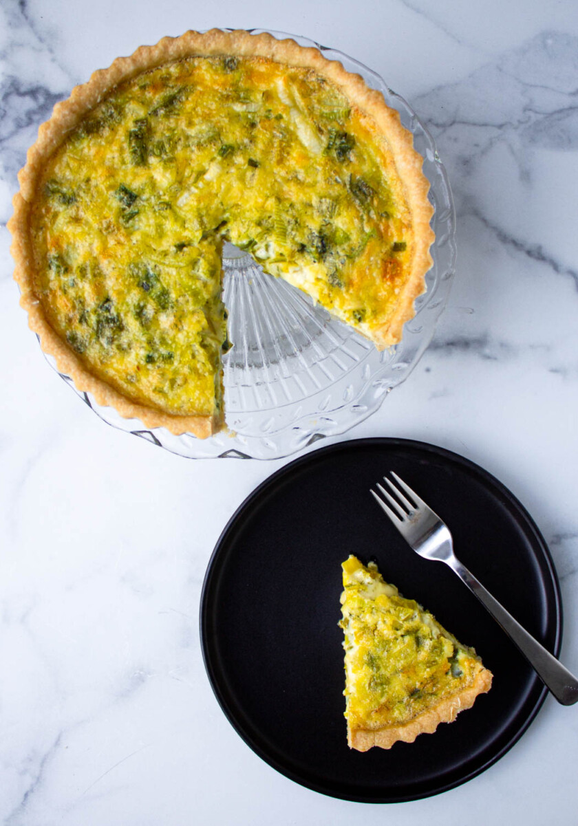 Una porción de quiche servida en un plato negro con un tenedor, junto al resto de la quiche en un molde para pastel de vidrio.
