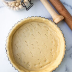 Unbaked pie crust in a fluted dish prepared for blind baking, alongside ceramic pie weights and a rolling pin on a marble surface.