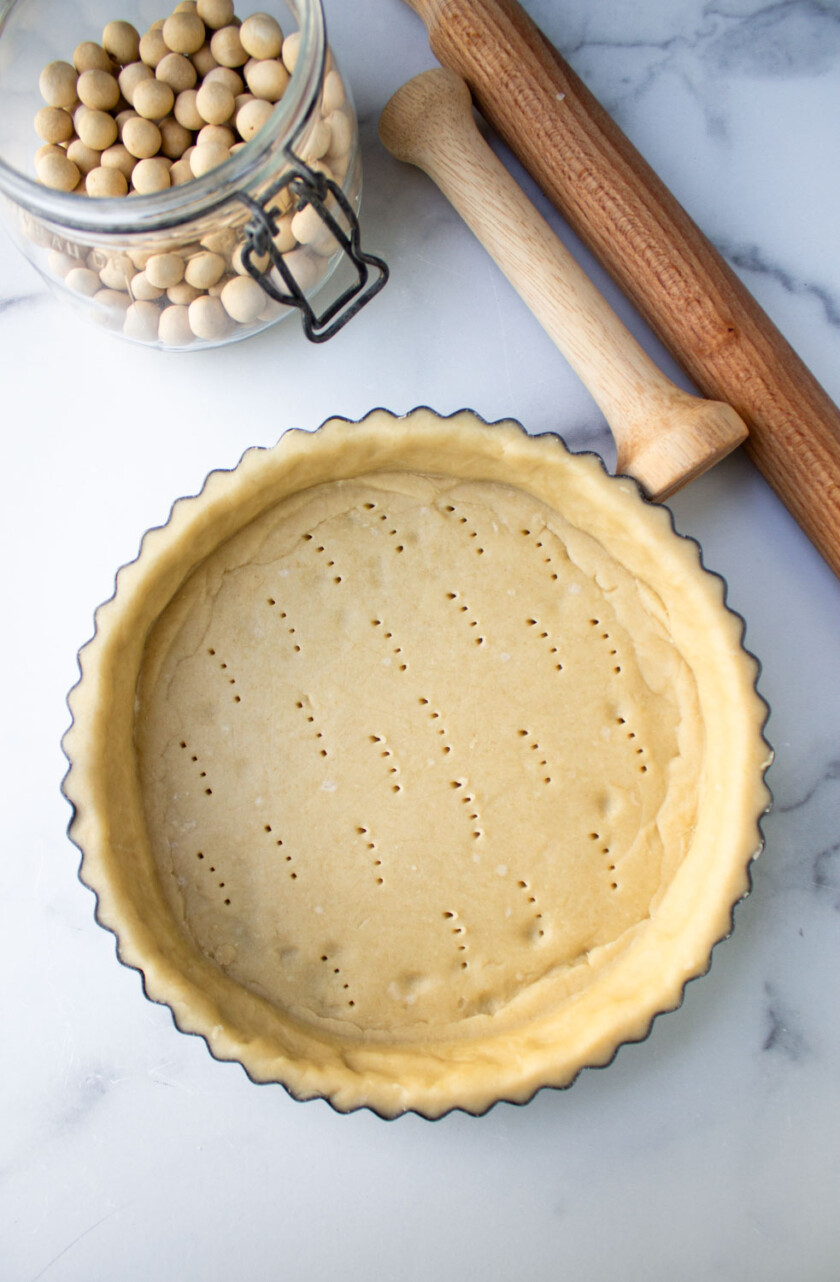 Unbaked pie crust in a fluted dish prepared for blind baking, alongside ceramic pie weights and a rolling pin on a marble surface.