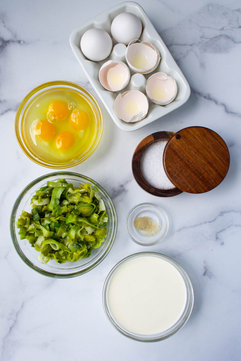 Ingredientes para cocinar dispuestos sobre una encimera de mármol, incluidos huevos, puerros picados y diversos condimentos.