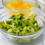 Chopped green leeks in a glass bowl with other cooking ingredients in the background.