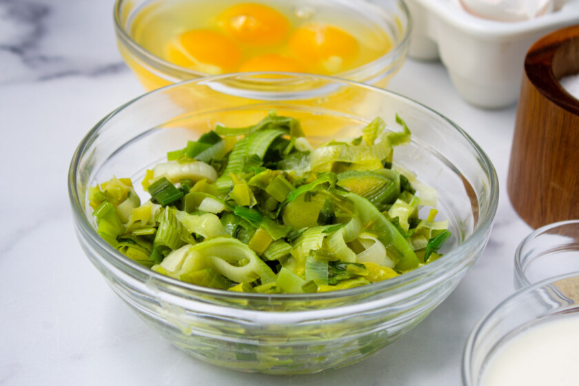 Puerros verdes picados en un recipiente de vidrio, con otros ingredientes para cocinar al fondo.