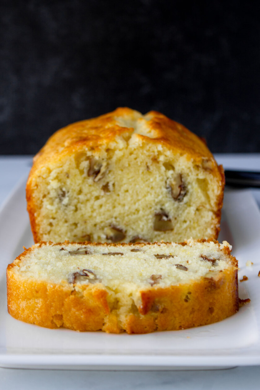 Un pan de limón con nueces, ya rebanado, sobre un plato blanco con un fondo oscuro.