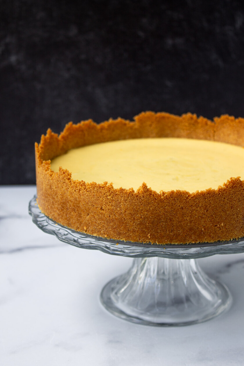 A whole key lime pie with a golden-brown crust on a glass stand, against a black and white marble background,