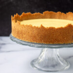 A creamy key lime pie with a golden crust on a glass stand against a marble countertop and dark background.