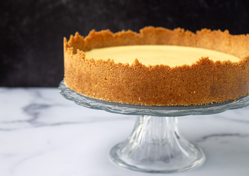 A creamy key lime pie with a golden crust on a glass stand against a marble countertop and dark background.
