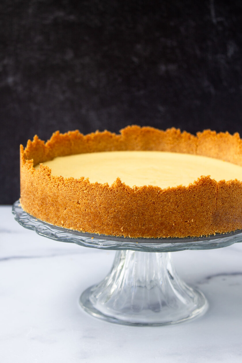 A creamy key lime pie with a golden-brown crust, presented on a clear glass stand against a white and dark gray backdrop.