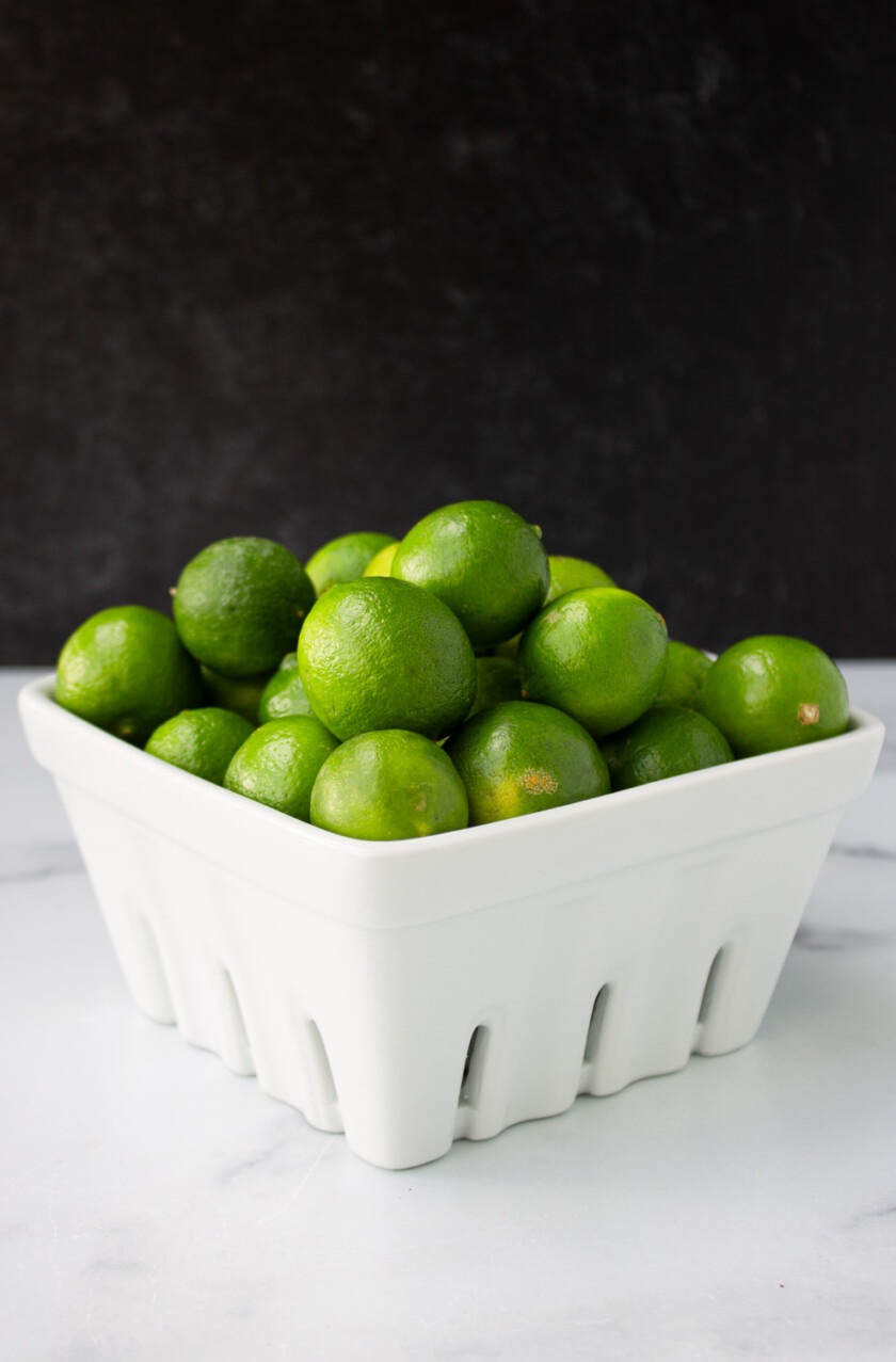 A white ceramic basket filled with fresh green limes on a marble surface against a dark background,
