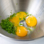 Tres huevos crudos con las yemas intactas y las hierbas verdes finamente picadas en un recipiente de acero inoxidable.