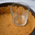 Vaso de vidrio transparente en un molde desmontable presionado con una capa de corteza de galleta Graham desmenuzada sobre una superficie de mármol.