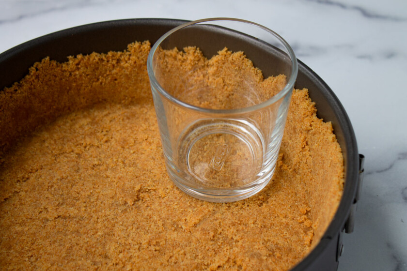 Glass inside a springform pan containing a pressed graham cracker crust, set on a marble countertop.