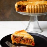 Un trozo de sabroso pastel de polvorosa de atún en un plato negro con un tenedor y el resto del pastel en un soporte al fondo.