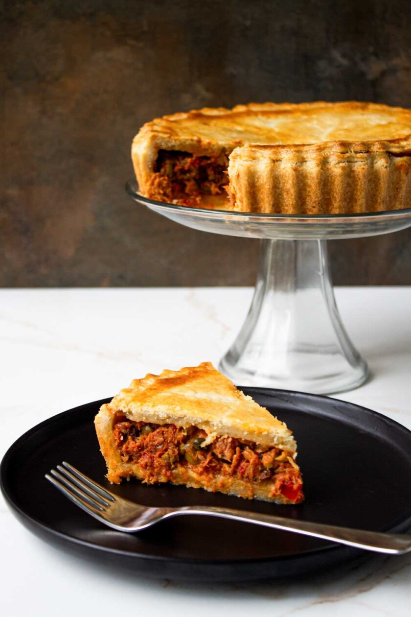 Un trozo de sabroso pastel de polvorosa de atún en un plato negro con un tenedor y el resto del pastel en un soporte al fondo.