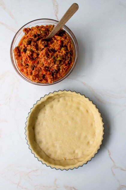 Un tazón de relleno de guiso de atún junto a una base de pastel sin hornear sobre una superficie de mármol.