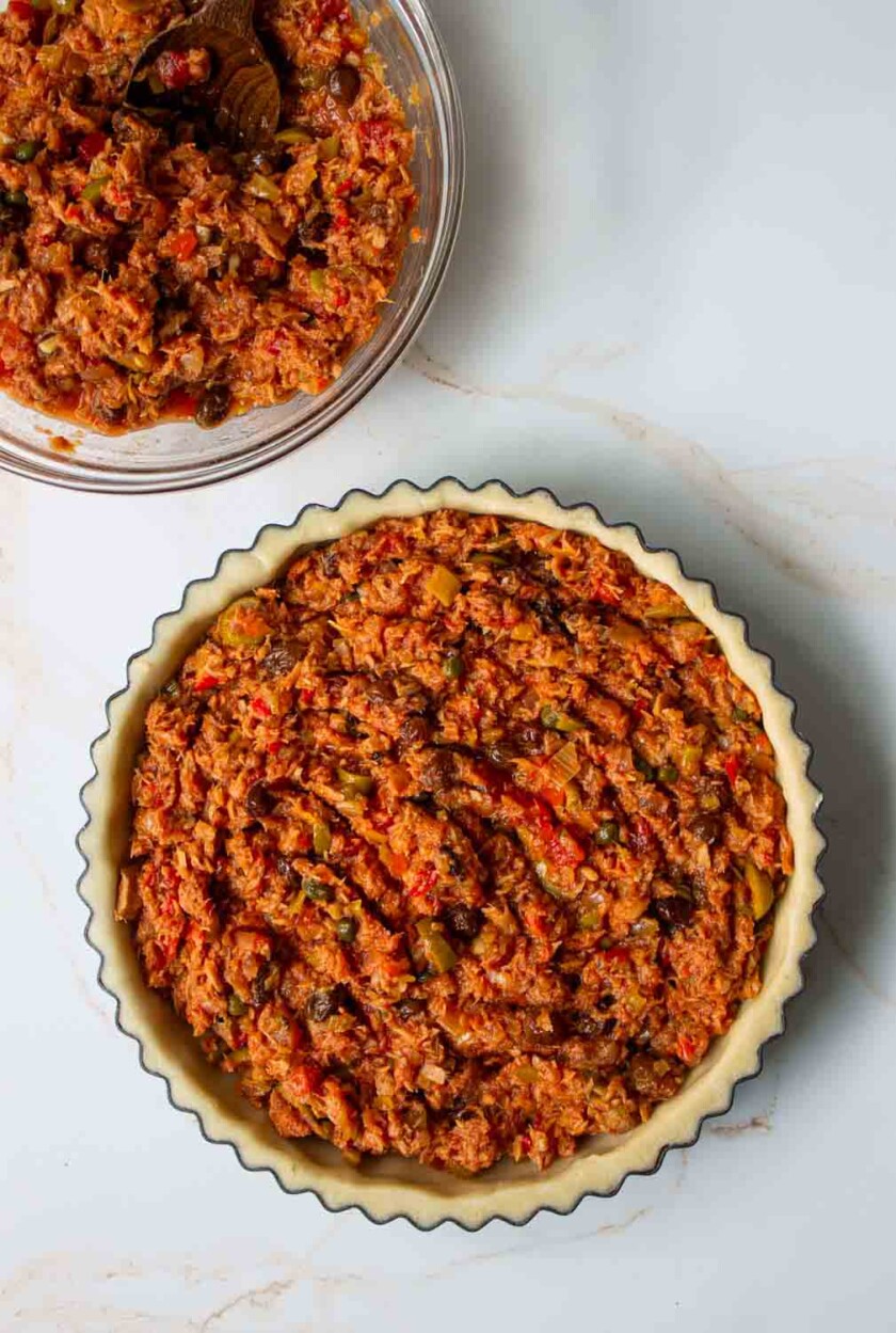 Vista aérea de un relleno de pastel de atún en un molde para tarta, junto a un tazón con relleno adicional. la mezcla parece densa con trozos visibles de verduras.