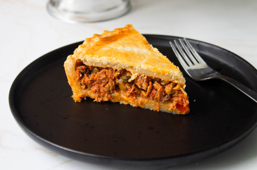 Un trozo de pastel de polvorosa de atún en un plato negro con un tenedor a un lado, mostrando una corteza hojaldrada y un relleno sabroso.