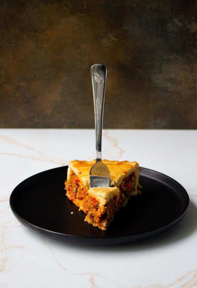 Un trozo de pastel de atún con corteza dorada, servido en un plato negro con un tenedor, sobre una encimera de mármol y un fondo gris texturizado.