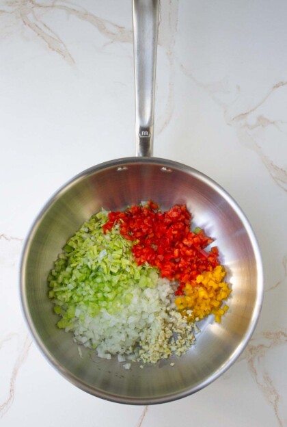Sartén de acero inoxidable con pimientos y cebollas verdes, amarillos y rojos finamente picados, dispuestos en secciones de colores separados sobre una encimera de mármol.
